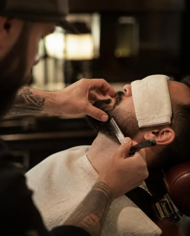 Barbero perfilando una barba a navaja, con una toalla sobre los ojos del cliente