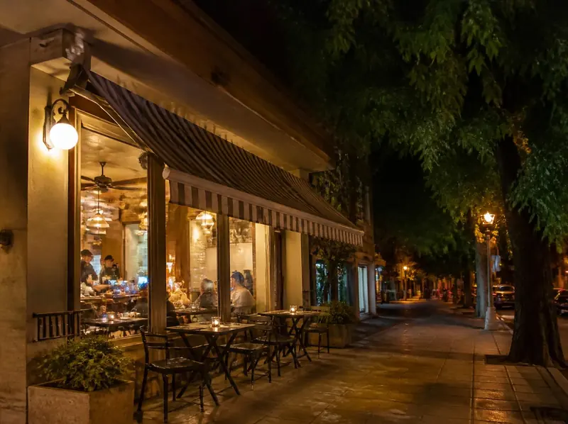 Frente de la pizzería de noche, con toldo a rayas, luces encendidas y mesas en la vereda