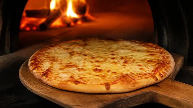 Pizza de muzzarella recién salida del horno a leña, sobre la pala de madera, con el fuego encendido atrás